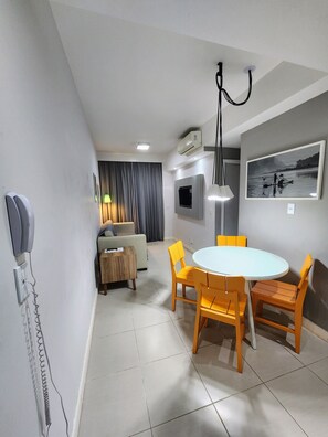A modern hotel room with a dining area featuring a round table and yellow chairs, a sofa, a TV, and an air conditioning unit.