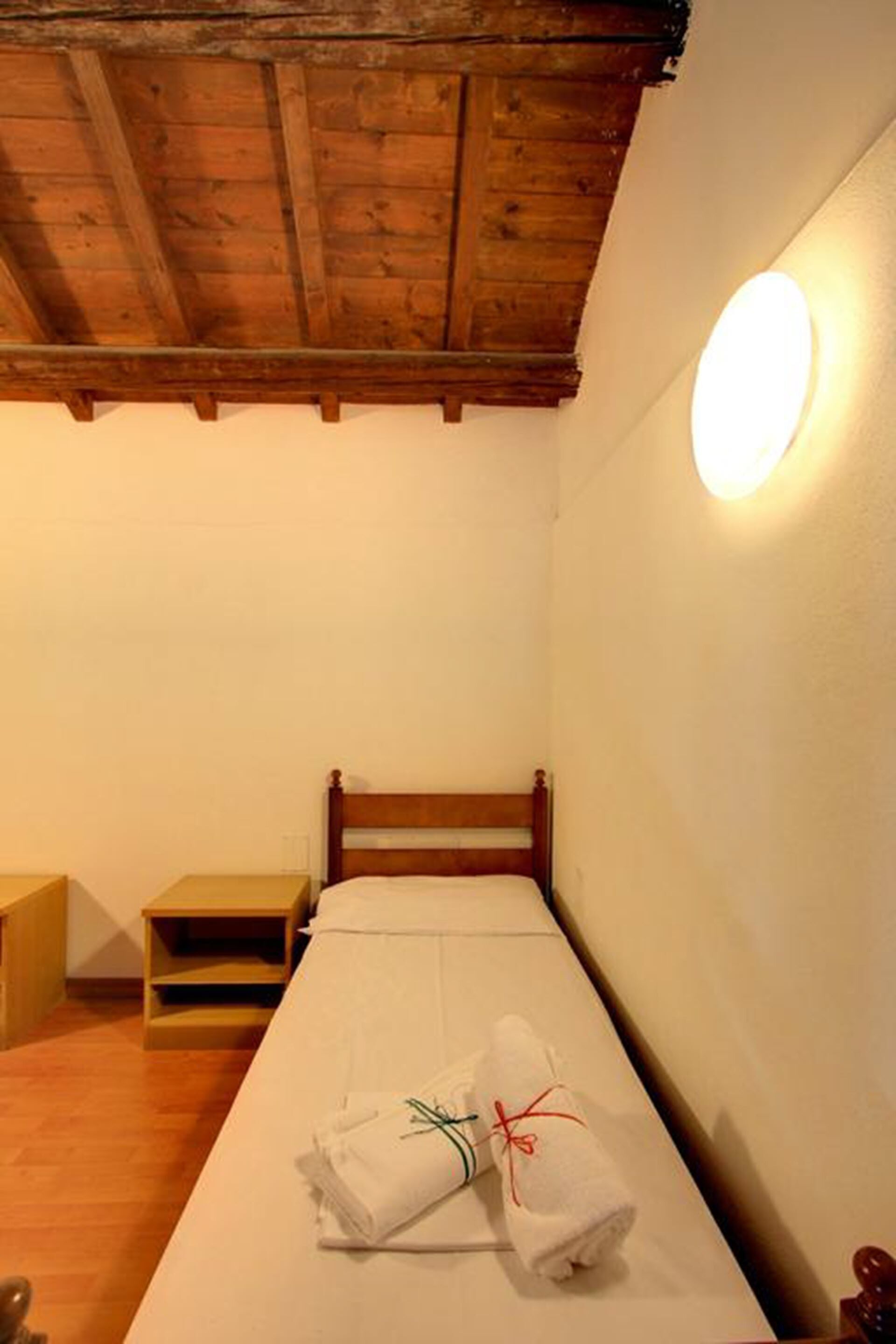 A small, single-bed room with a wooden ceiling and a round wall light.