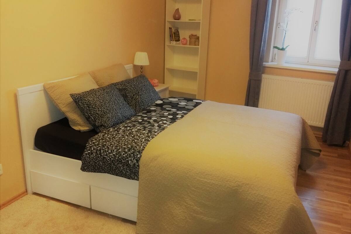 Un dormitorio con cama, almohadas, una mesita de noche, lámpara, estante con objetos decorativos y ventana con cortinas.