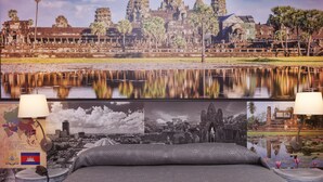 Una cama con una colcha estampada, dos lámparas de noche y una pared decorada con una fotografía grande de Angkor Wat y una fotografía más pequeña de un templo.