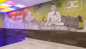 Vestíbulo de hotel con un mural que muestra una estatua de Buda y el Taj Mahal, un letrero que dice 'un estilo diferente para dormir' y el nombre del hotel 'JC HOTELS'.