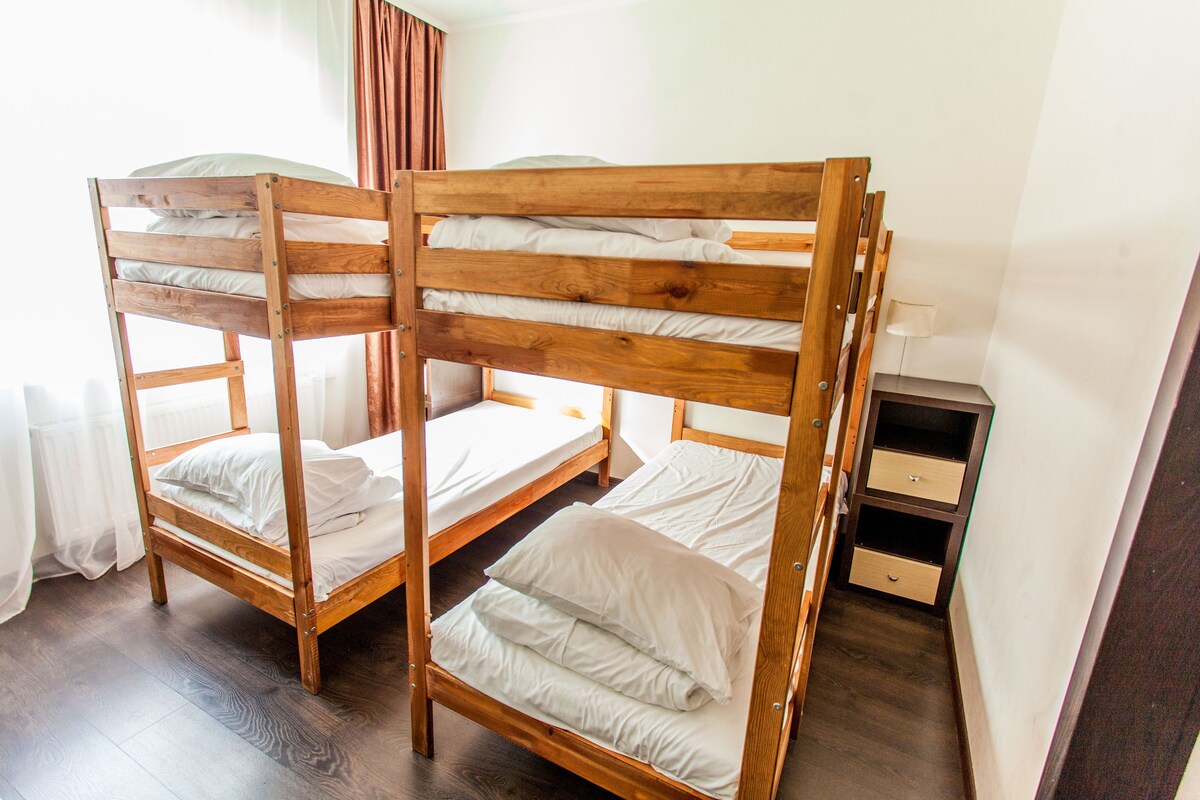 A room with three wooden bunk beds, a small bedside table with drawers, and a window with curtains.