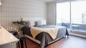 Une chambre moderne avec un grand lit, une table de chevet, une plante en pot et une vue sur la ville par une grande fenêtre.
