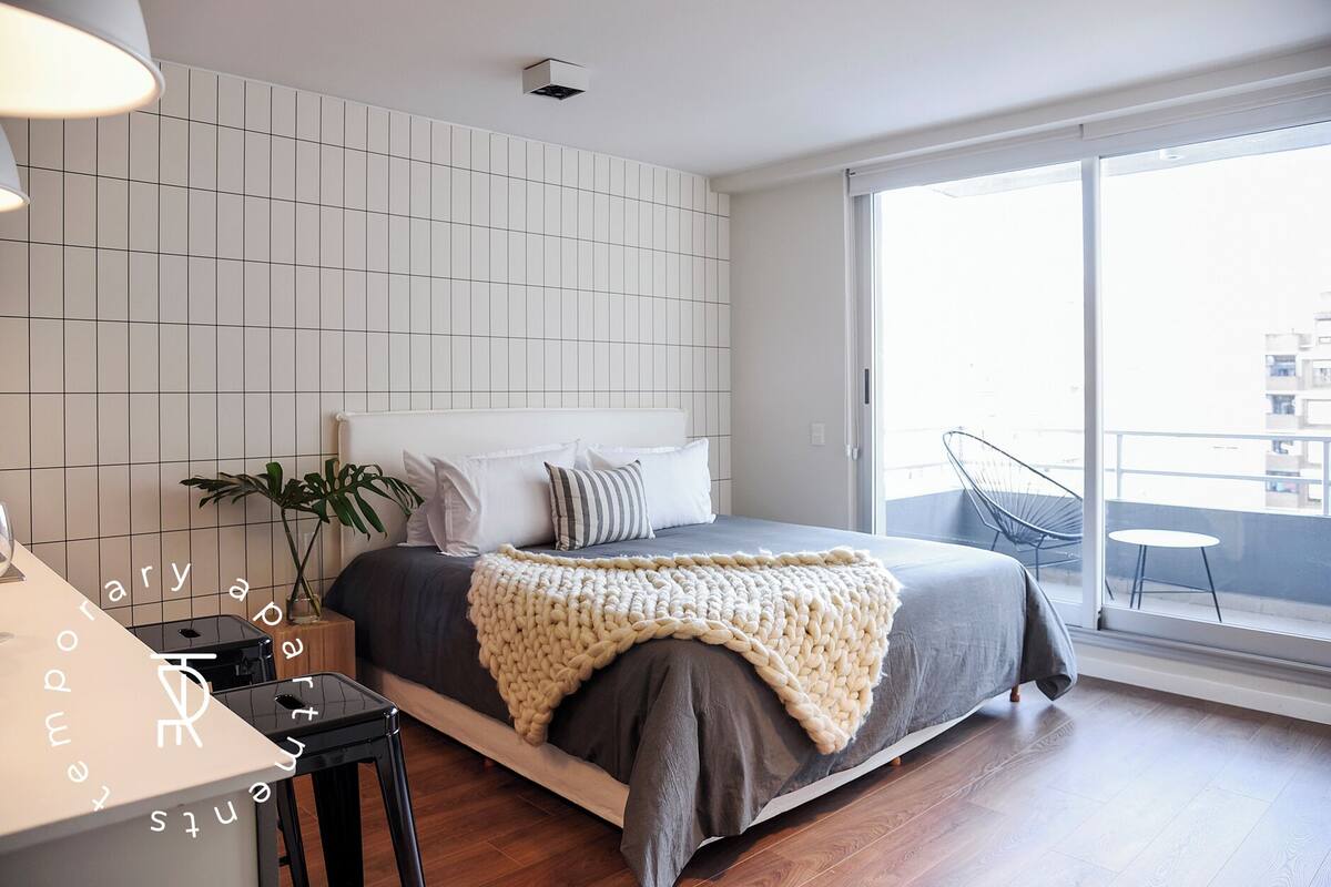 A modern bedroom with a large bed, a bedside table, a potted plant, and a view of the city through a large window.