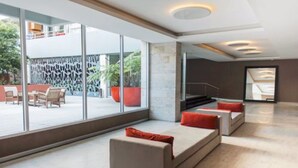 Un hall d’hôtel moderne avec un grand mur de verre, un espace salon doté de canapés et de coussins rouges, et un escalier menant à un niveau supérieur.
