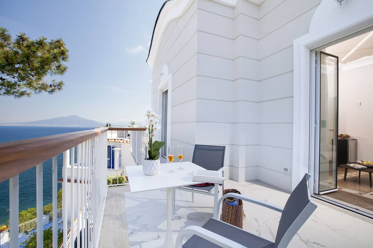 Un balcon avec une table blanche, deux chaises, une plante en pot et une vue sur la mer et une montagne.