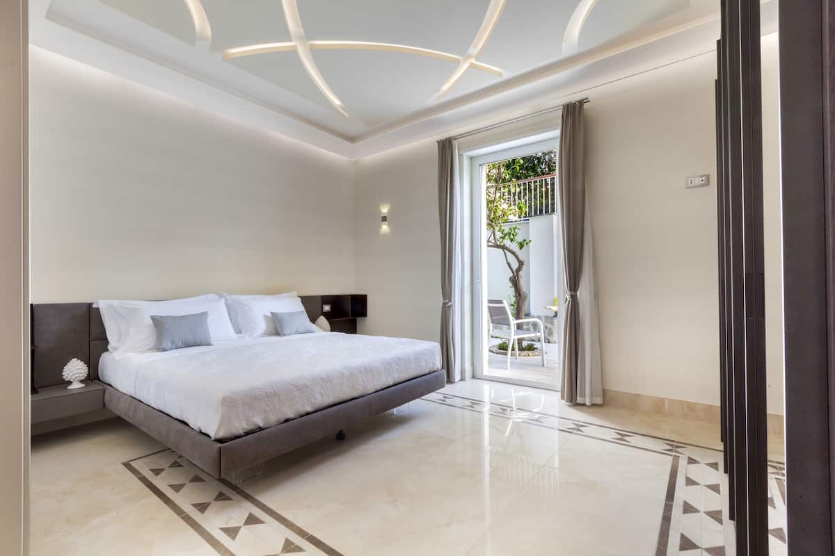 Une chambre moderne avec un grand lit, une table de chevet, une lampe fixée au mur et une vue sur un espace extérieur grâce à une porte vitrée.