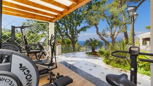 Un área de gimnasio al aire libre con equipo de ejercicio, un pérgola de madera y vistas al mar.