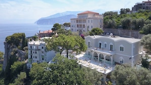 Un edificio costero con balcones que dan al mar, rodeado de vegetación y otros edificios.