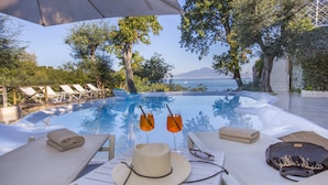 Escena junto a la piscina con sillones de descanso, una mesa con un sombrero y gafas, y vistas al mar y a la montaña.
