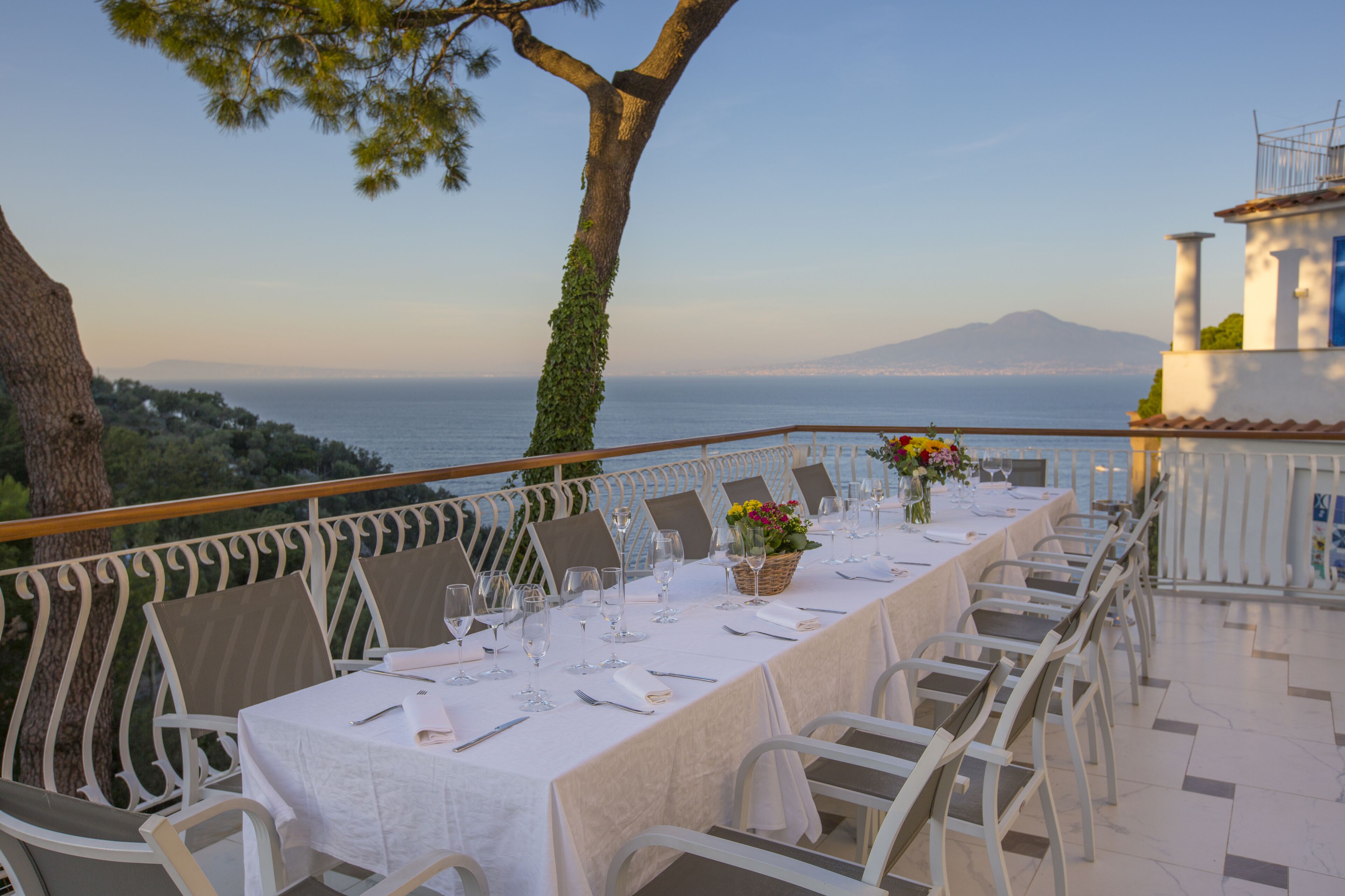 A dining table set for a meal with white tablecloths and chairs, overlooking a scenic view of the sea and a mountain.