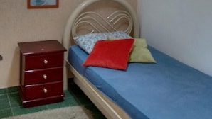 Une petite chambre avec un lit simple, une tête de lit en bois, une commode rouge, un miroir et une fenêtre avec des stores.