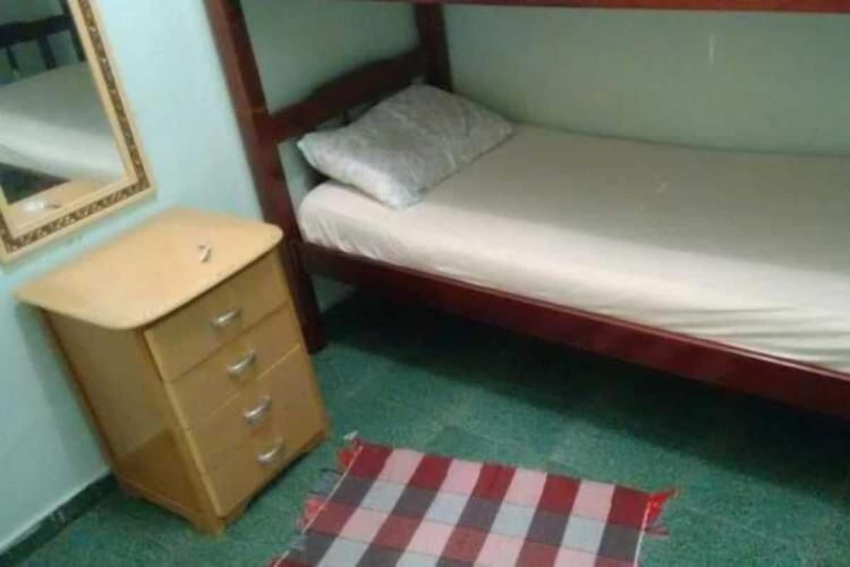Une petite chambre avec un lit simple, une tête de lit en bois, une table de chevet avec des tiroirs, un miroir et un tapis à carreaux sur le sol.