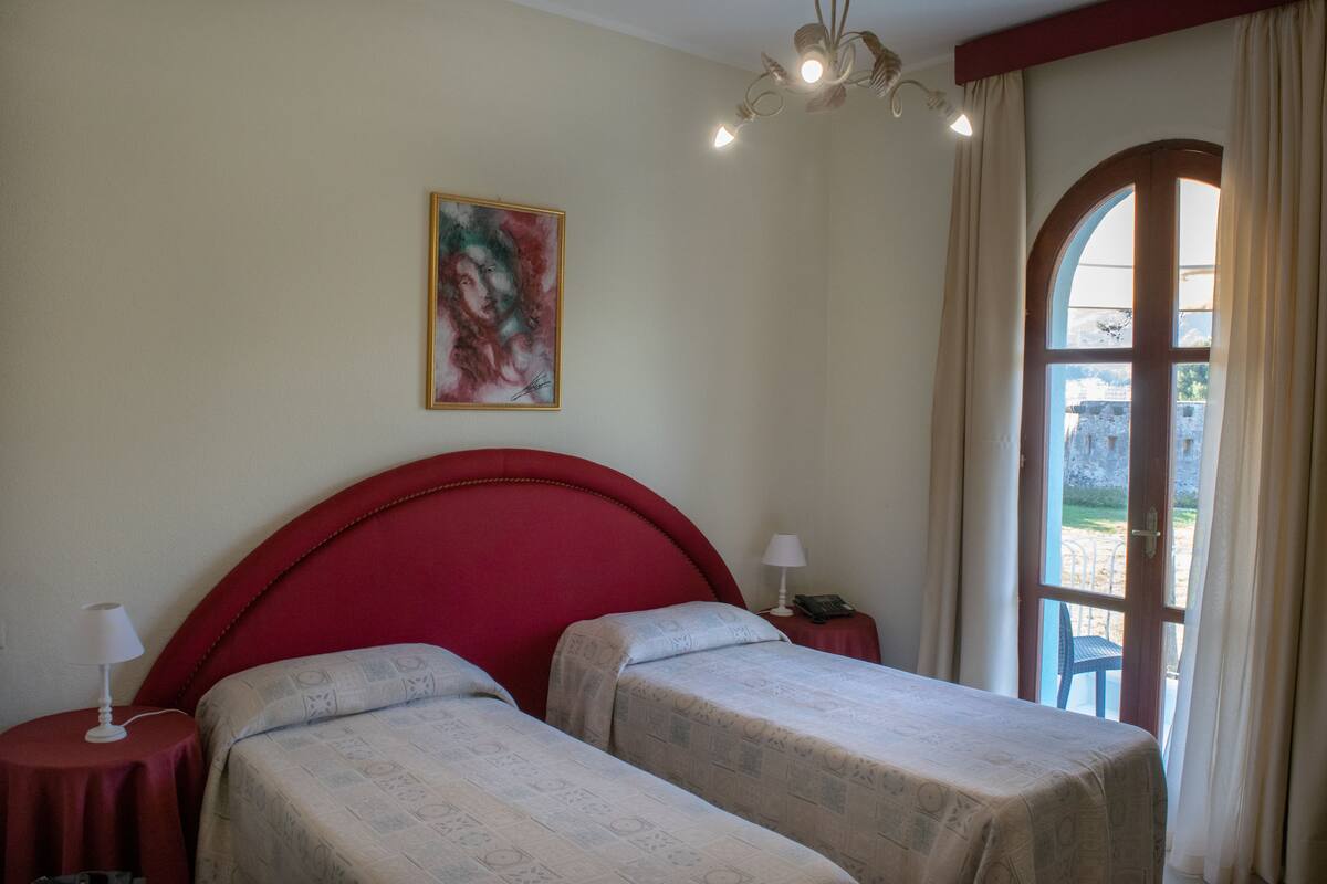 Une chambre avec une tête de lit rouge, deux lits avec une literie à motifs, une petite table de chevet avec une lampe, un tableau au mur et une fenêtre avec des rideaux.