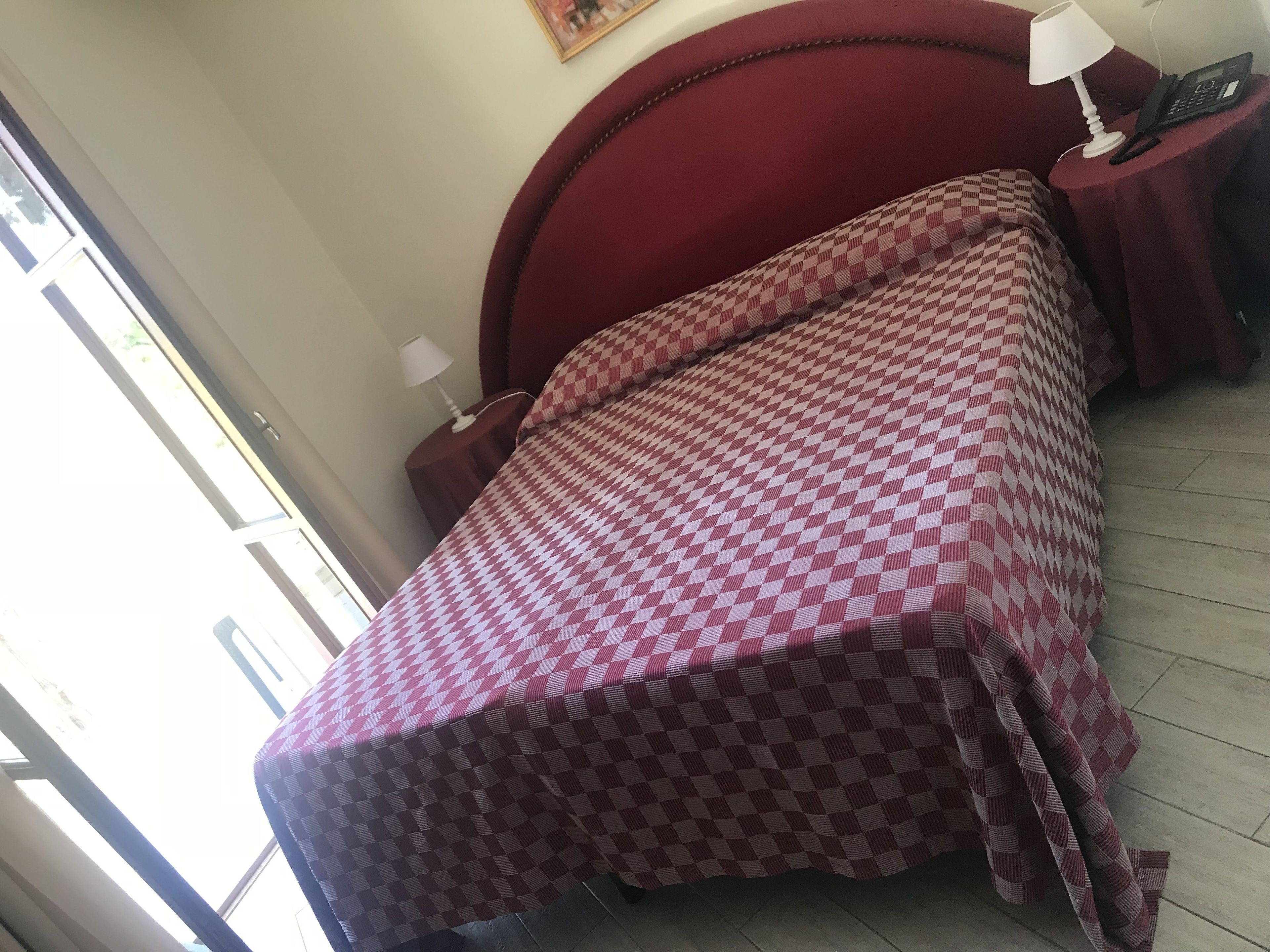 Een kamer met een geruit beddengoed, een rode bekleden hoofdeinde, een nachtkastje met een lamp en een telefoon, en een raam met uitzicht op buiten.