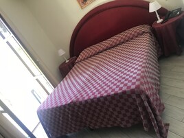 Una habitación con colcha a cuadros, cabecero tapizado en rojo, mesita de noche con lámpara y teléfono, y ventana con vistas al exterior.