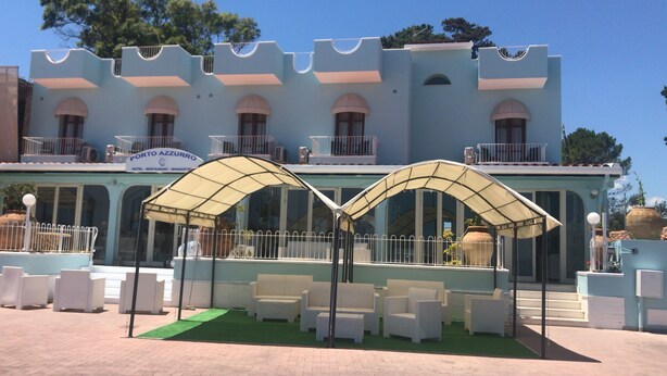 Een hotel met een lichtblauwe gevel, een terras met witte meubels en een bord waarop staat: "Porto Azzurro Hotel - Restaurant - Banquet".
