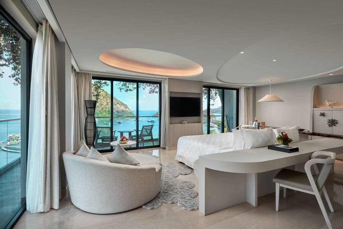 Une chambre d’hôtel moderne dotée d’une grande fenêtre donnant sur la mer, d’un coin salon confortable, d’un lit et d’une table à manger.