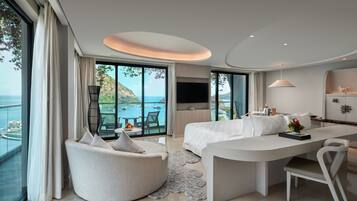 A modern hotel room with a large window offering a view of the sea, a cozy seating area, a bed, and a dining table.