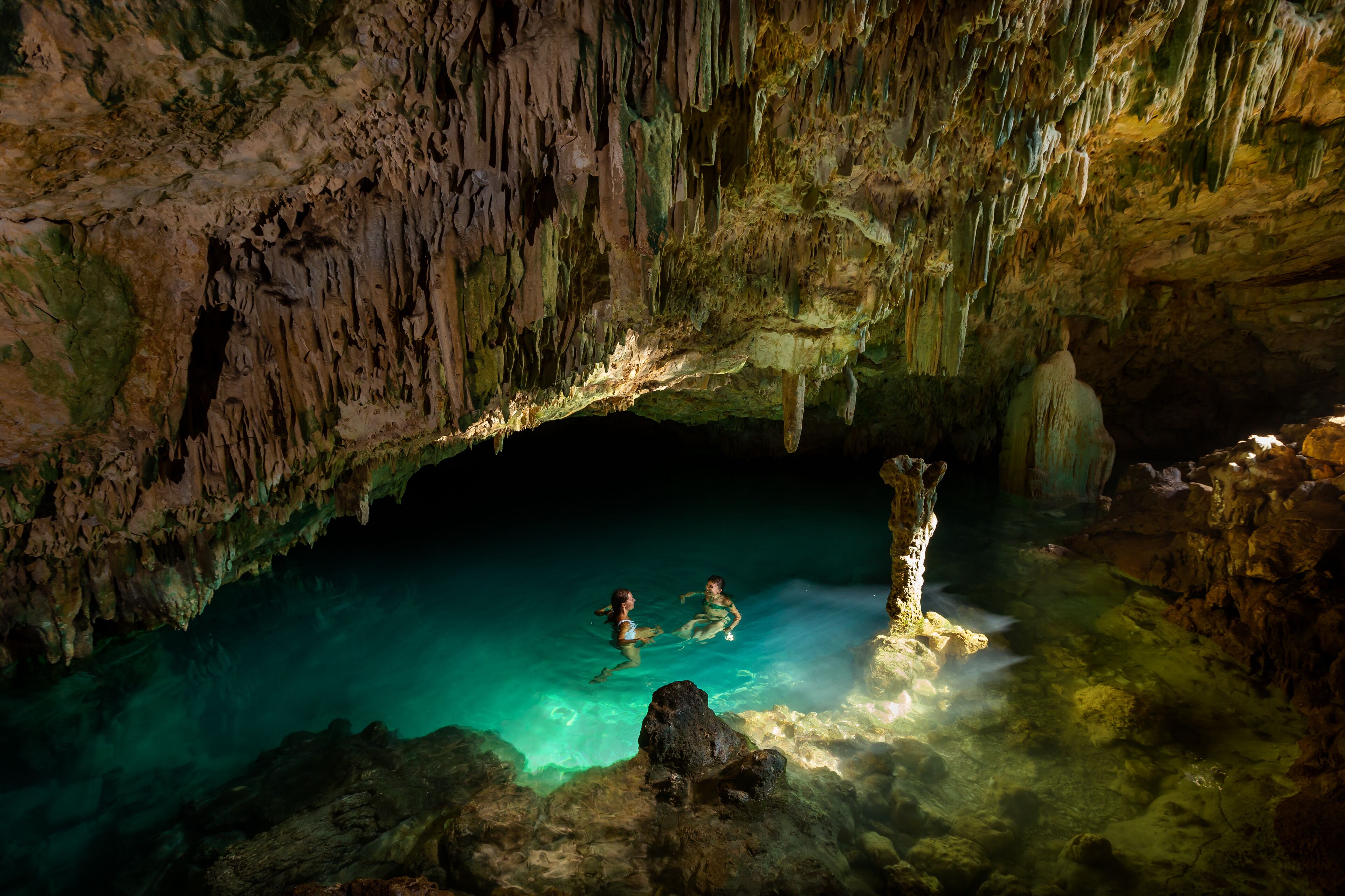 Eine Höhle mit Stalaktiten und Stalagmiten, klarem Wasser und zwei darin schwimmenden Personen.