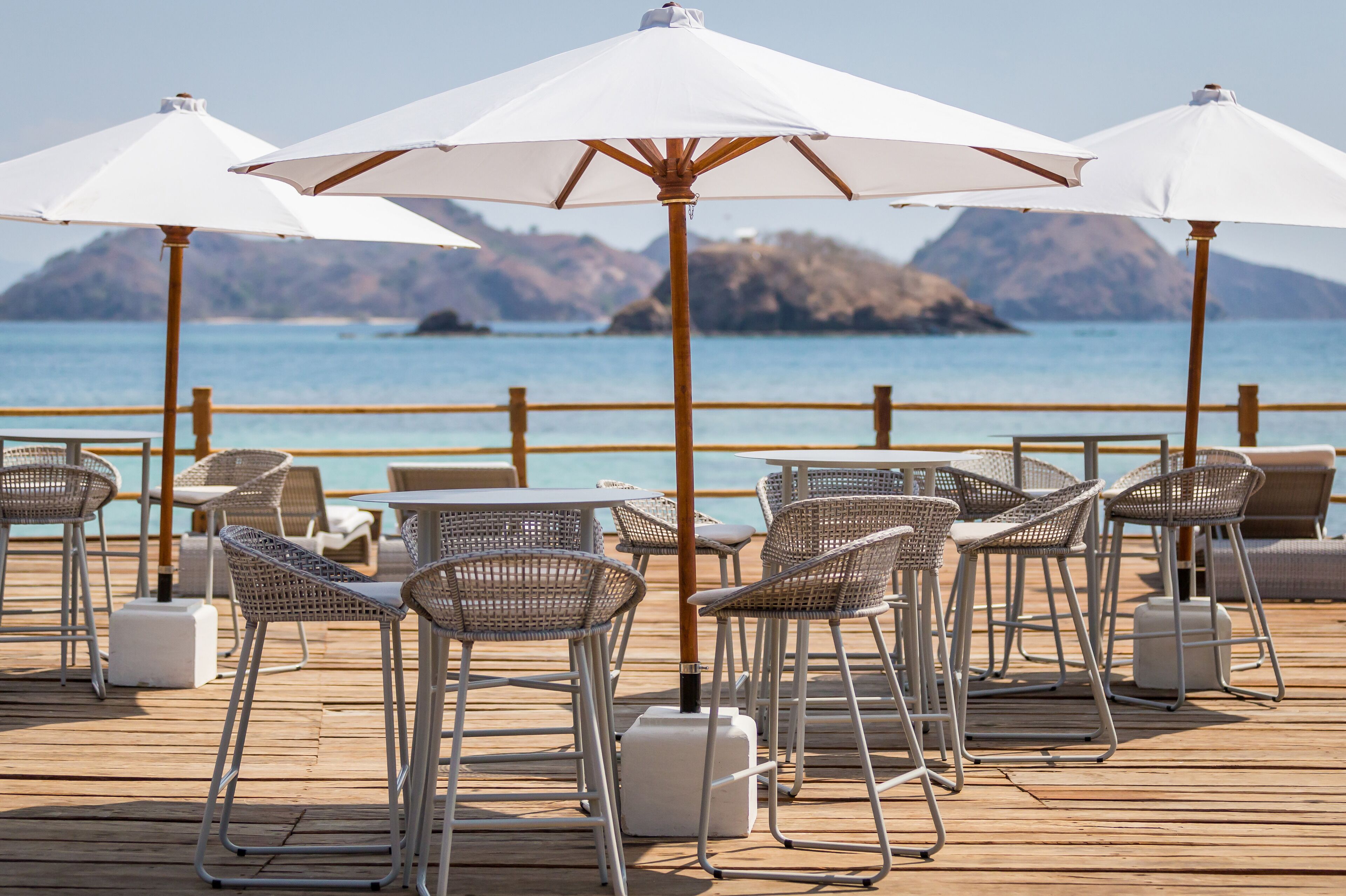 A deck with wicker chairs and white umbrellas, overlooking a body of water with mountains in the background.
