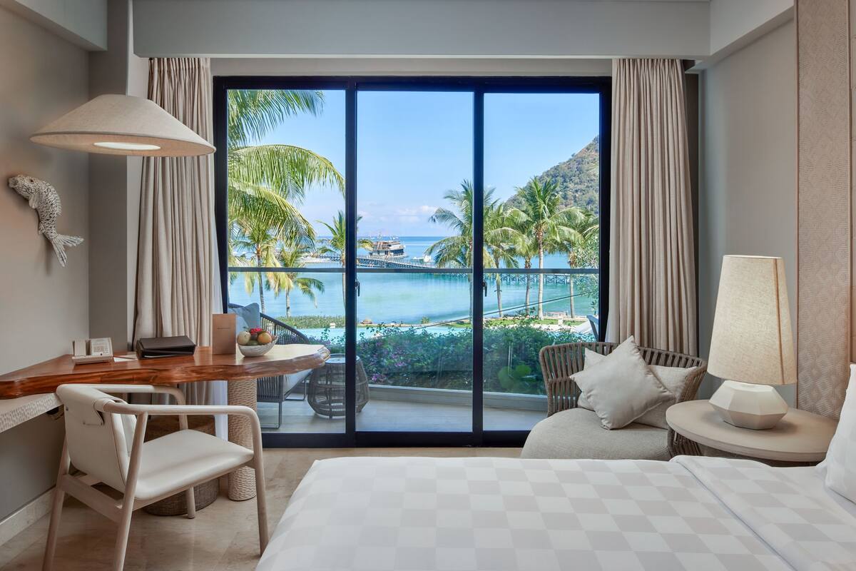 Une chambre d’hôtel avec une grande fenêtre, un bureau avec une chaise, un lit et une lampe.