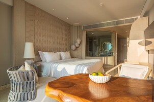 A hotel room with a bed, a wooden table with a bowl of fruit, a chair, and a lamp.