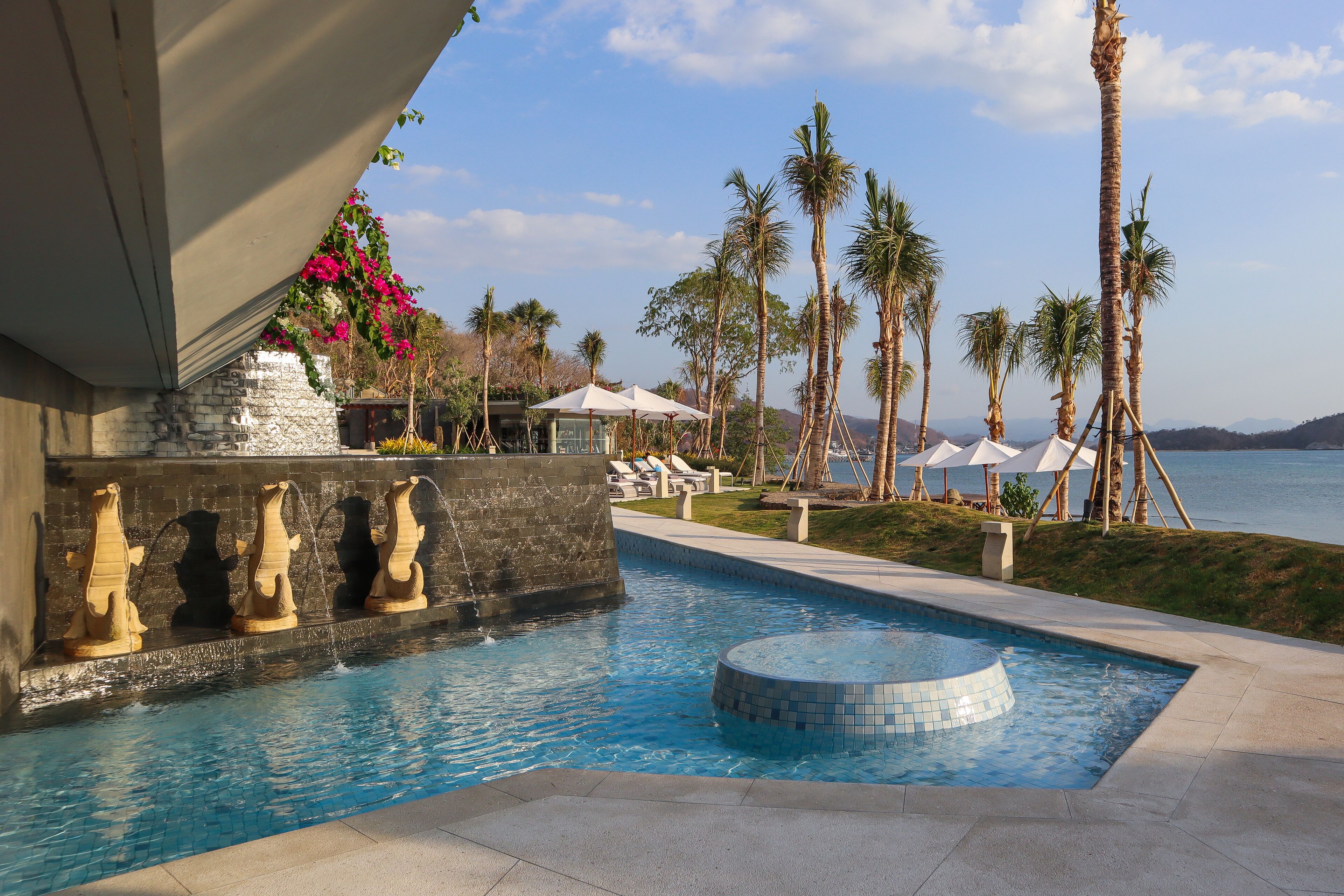 A swimming pool with a water feature, surrounded by palm trees and lounge chairs.