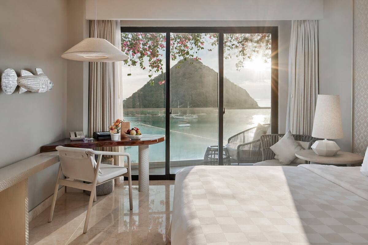 Une chambre d’hôtel avec un lit, un bureau, une chaise, une table avec des fruits, et une vue sur une montagne et un plan d’eau.