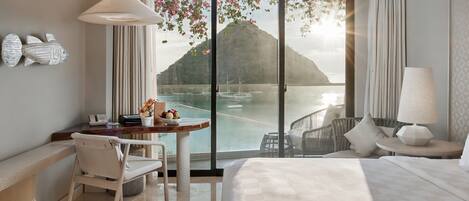 Habitación de hotel con cama, escritorio, silla, mesa con frutas y vista a una montaña y al agua.
