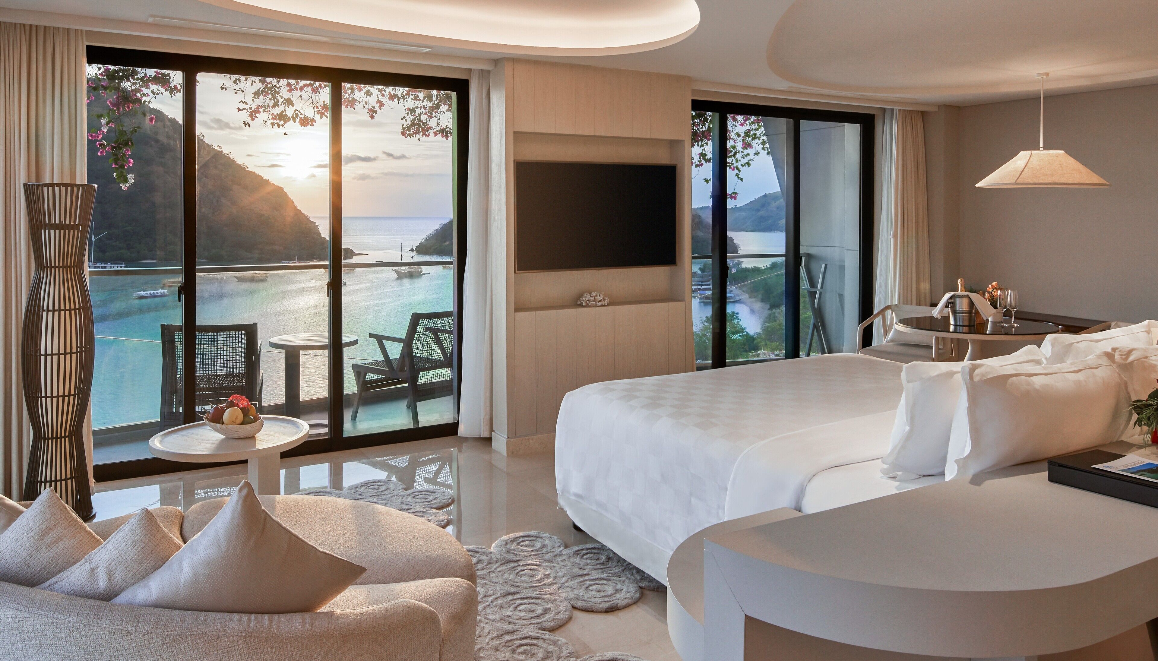 A hotel room with a large bed, a TV, a balcony with a table and chairs, and a view of the sea.