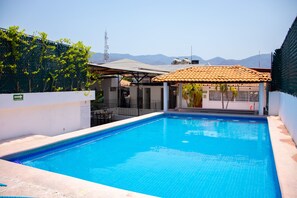 Una piscina rectangular con agua azul cristalina, rodeada por un muro blanco y una valla verde. Se observa un edificio con techo de tejas y un cartel que indica restaurante o cafetería.