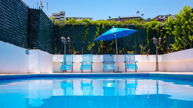 Una piscina con agua azul cristalina, rodeada de paredes blancas y una valla verde. Varias sillas azules y una sombrilla azul ofrecen sombra.