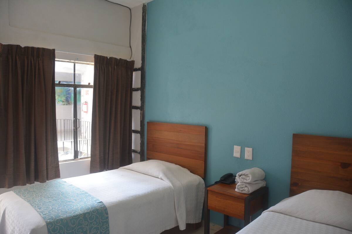 A hotel room with two single beds, a wooden headboard, a small bedside table with a phone and towels, and a window with curtains.