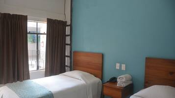 Habitación de hotel con dos camas individuales, un cabecero de madera, una mesita de noche pequeña con un teléfono y toallas, y una ventana con cortinas.