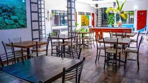 Un amplio comedor interior con múltiples mesas y sillas de madera, una escalera que conduce a un nivel superior, y varias plantas y obras de arte en las paredes.