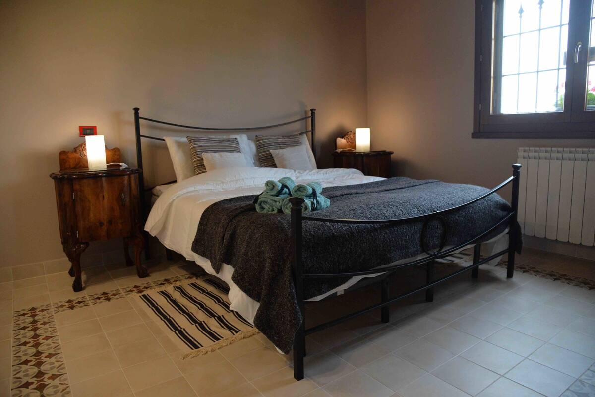A neatly made bed with a dark headboard, two bedside tables with lamps, a patterned rug, and a window with a view.