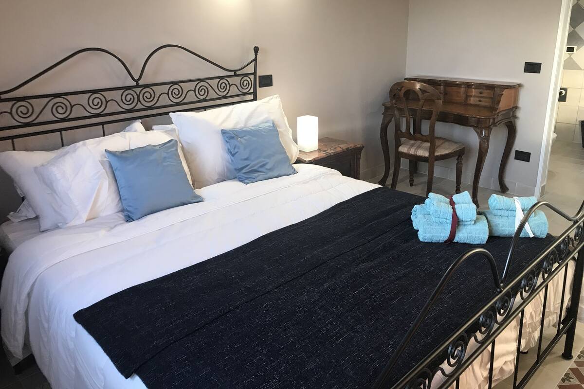 A neatly made bed with a black bedspread, white and blue pillows, and a wooden headboard. There is a small wooden desk with a chair beside the bed, and a lamp on the nightstand.