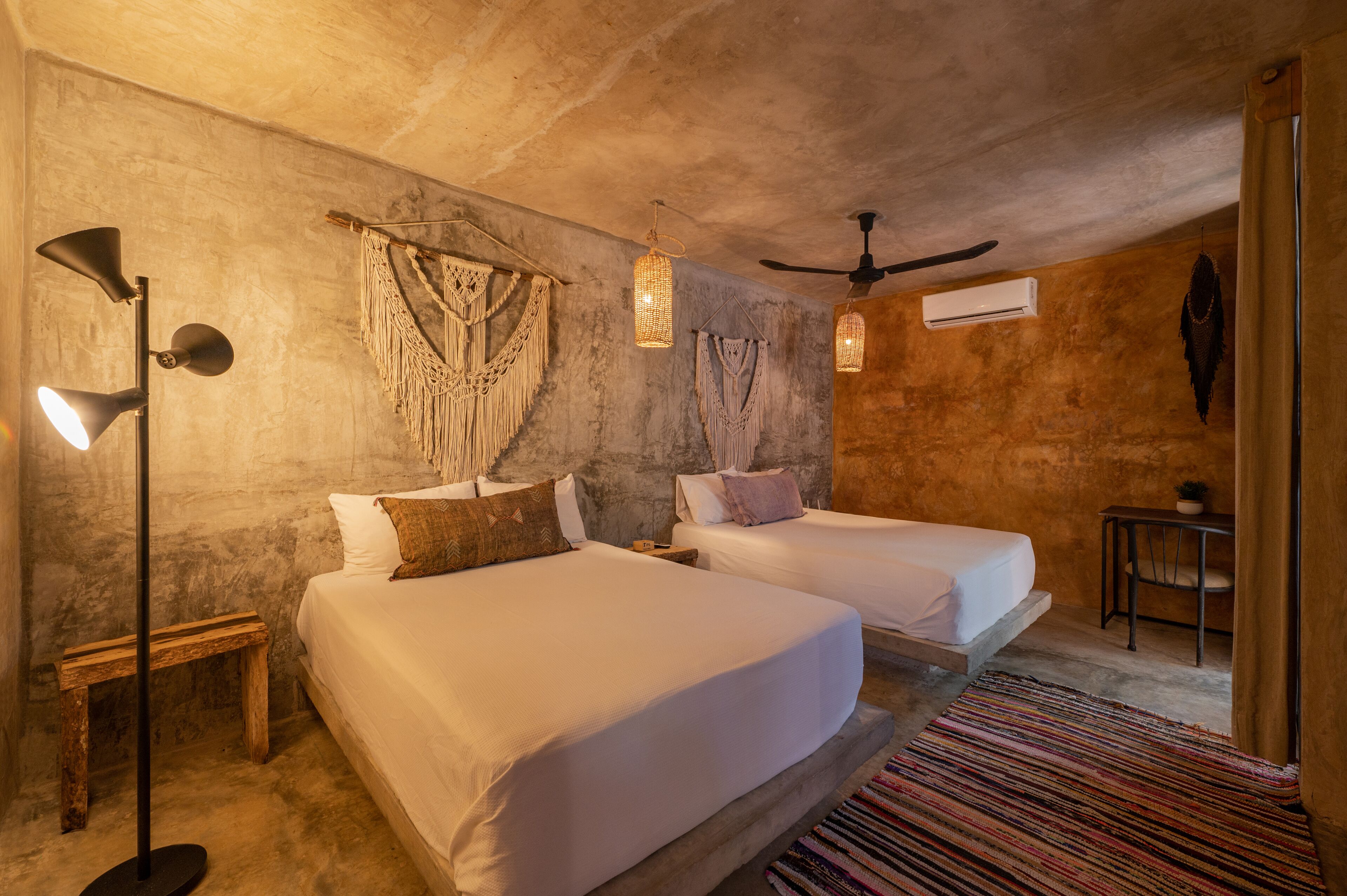 Una habitación con dos camas, ventilador de techo, macramé en las paredes y una pequeña mesa con una planta.