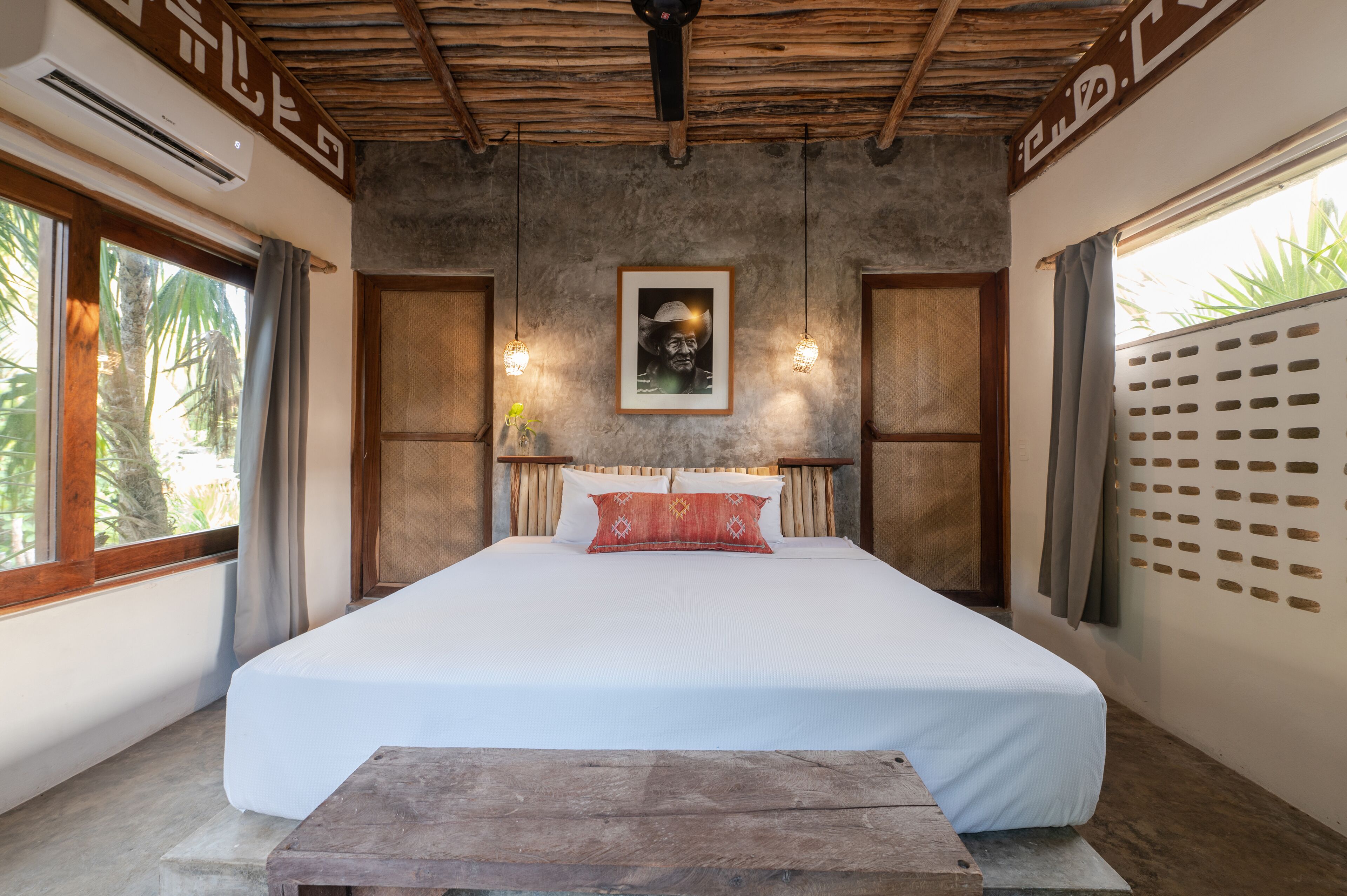 Una cama grande con cabecera de madera, un cuadro enmarcado encima y una ventana con cortinas a la izquierda.