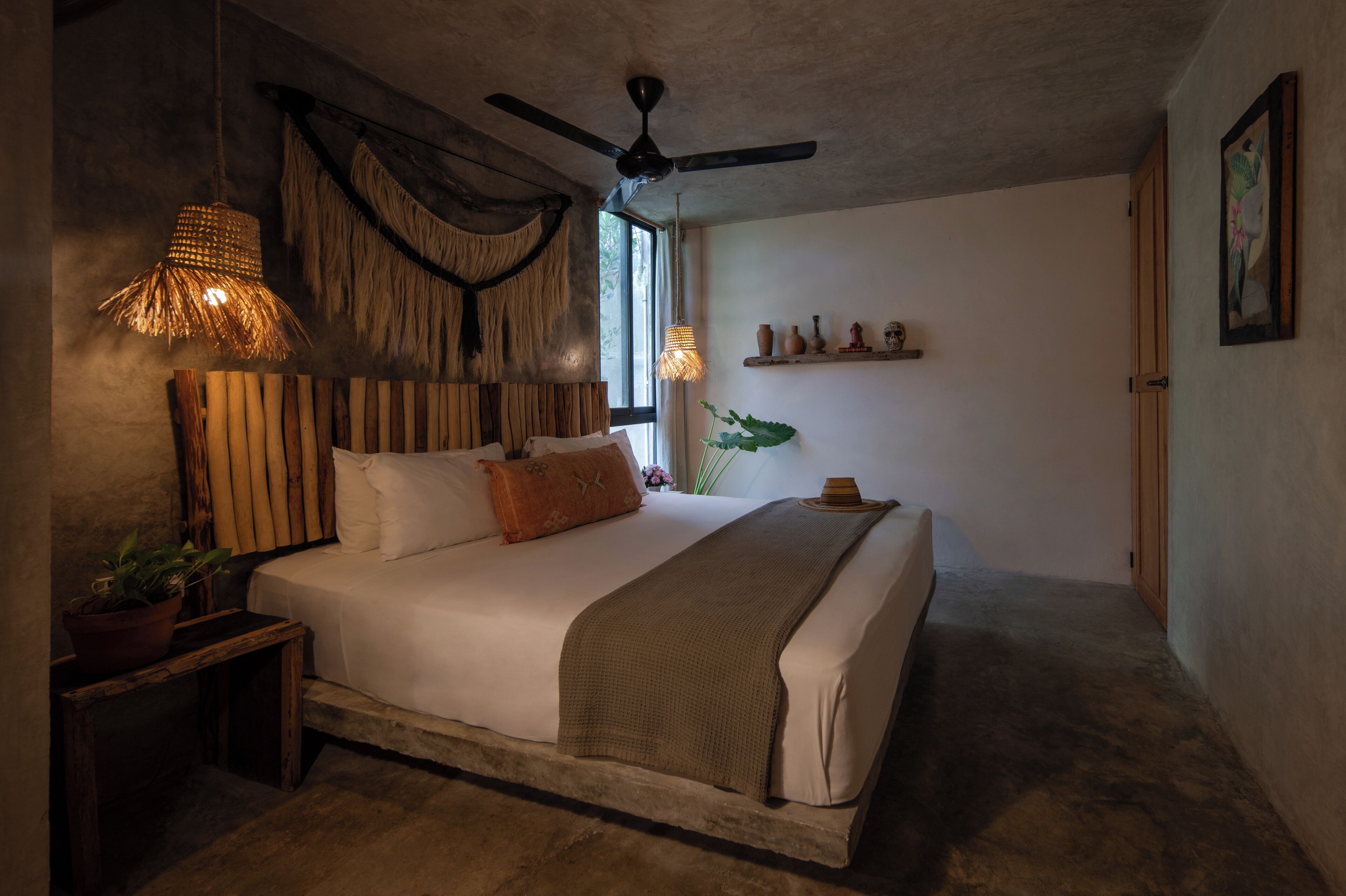 A bedroom with a large bed, a ceiling fan, a window with natural light, a plant, and a shelf with decorative items.