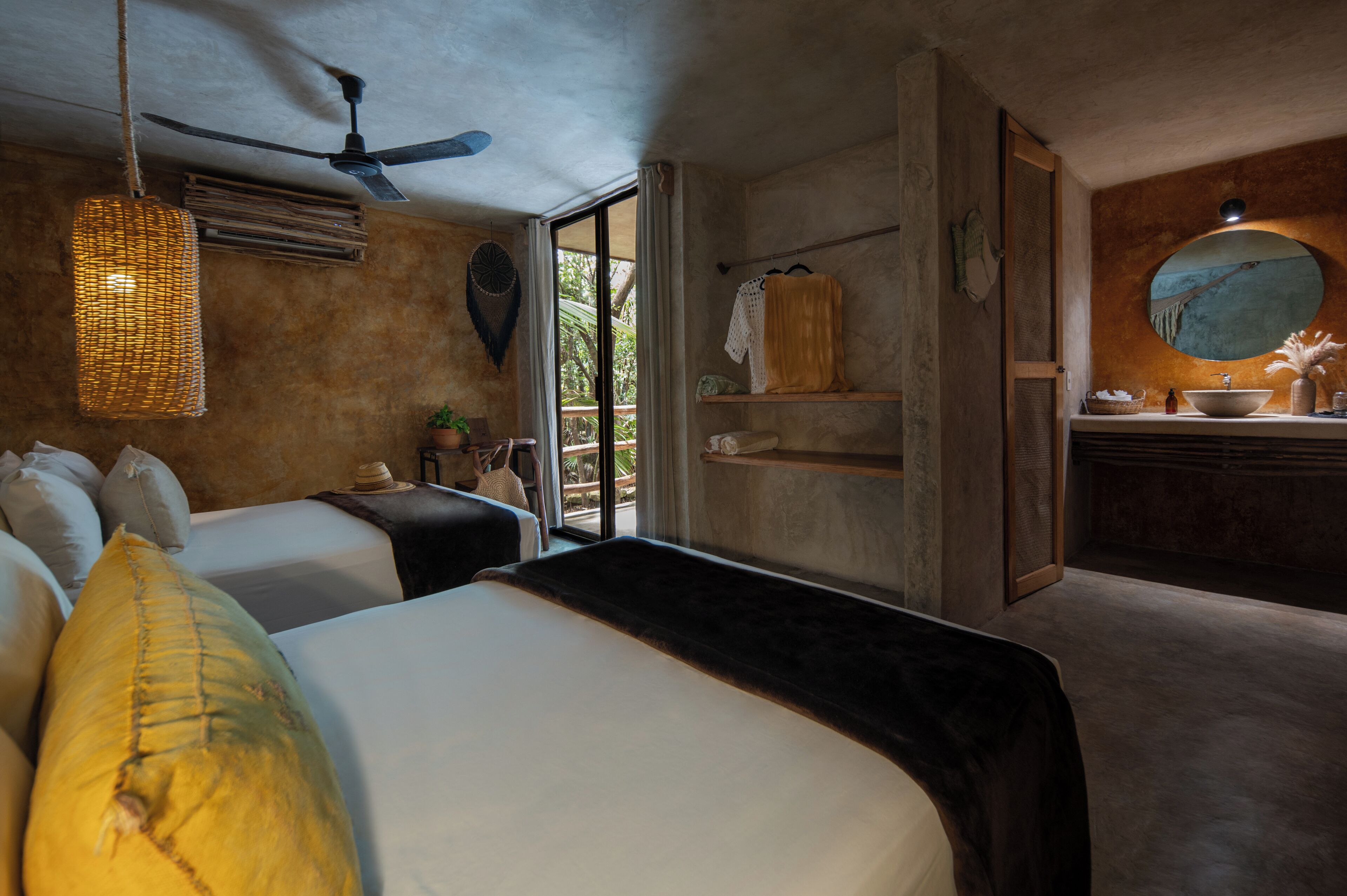 Un dormitorio con dos camas, ventilador de techo, una ventana grande y un baño con un espejo redondo y lavamanos.
