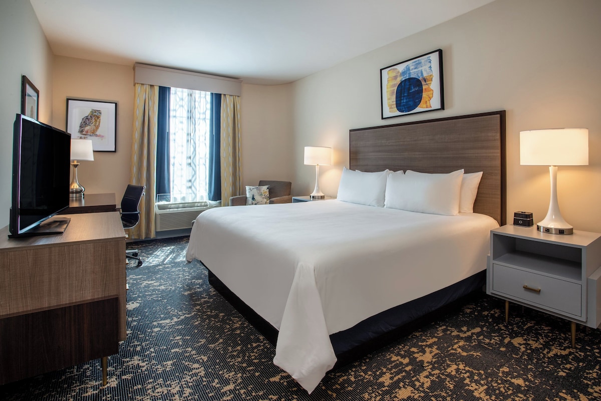 A hotel room with a large bed, two bedside tables, a desk, a television, and a window with curtains.