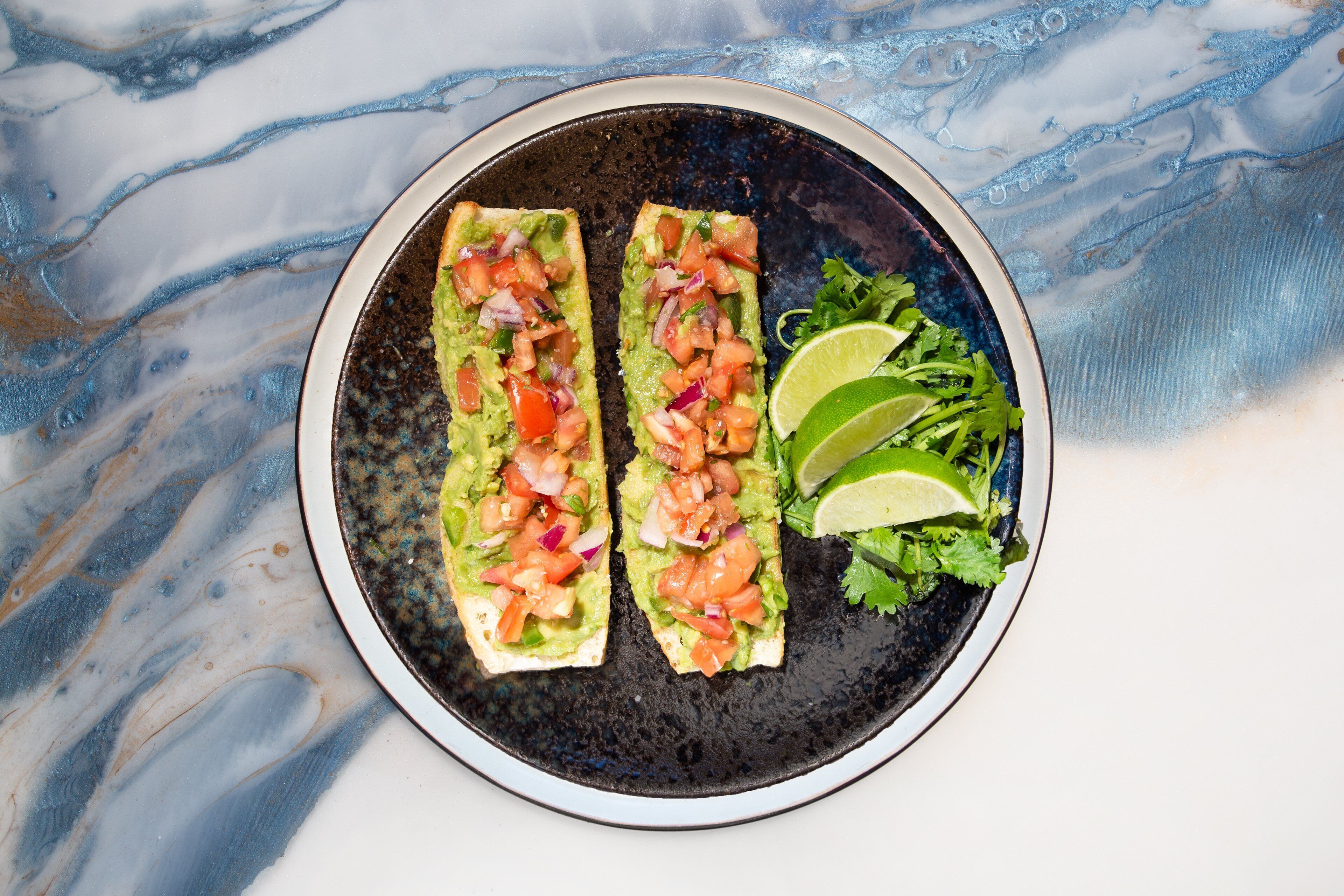 Zwei Avocado-Toast-Sandwiches auf einem Teller, dazu ein Limettenschnitz und ein kleiner Salat.