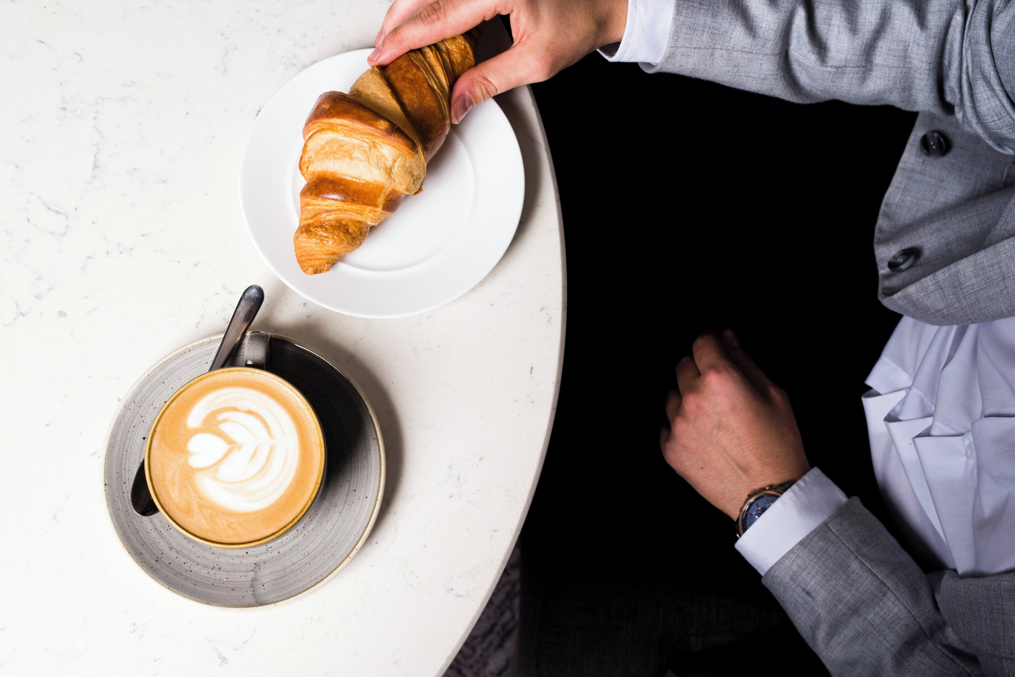Eine Person in einem grauen Anzug sitzt an einem Tisch mit einem Croissant und einer Kaffeetasse.