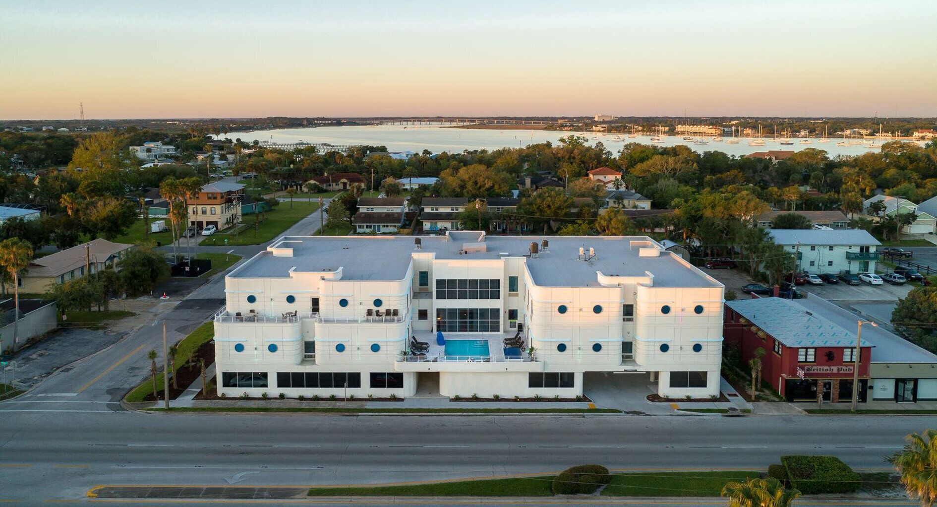 A modern building with a swimming pool, surrounded by a residential area and a body of water.