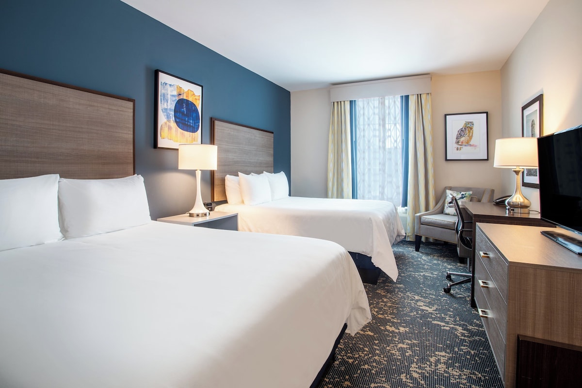 A hotel room with two beds, a desk, a chair, a television, and artwork on the walls.