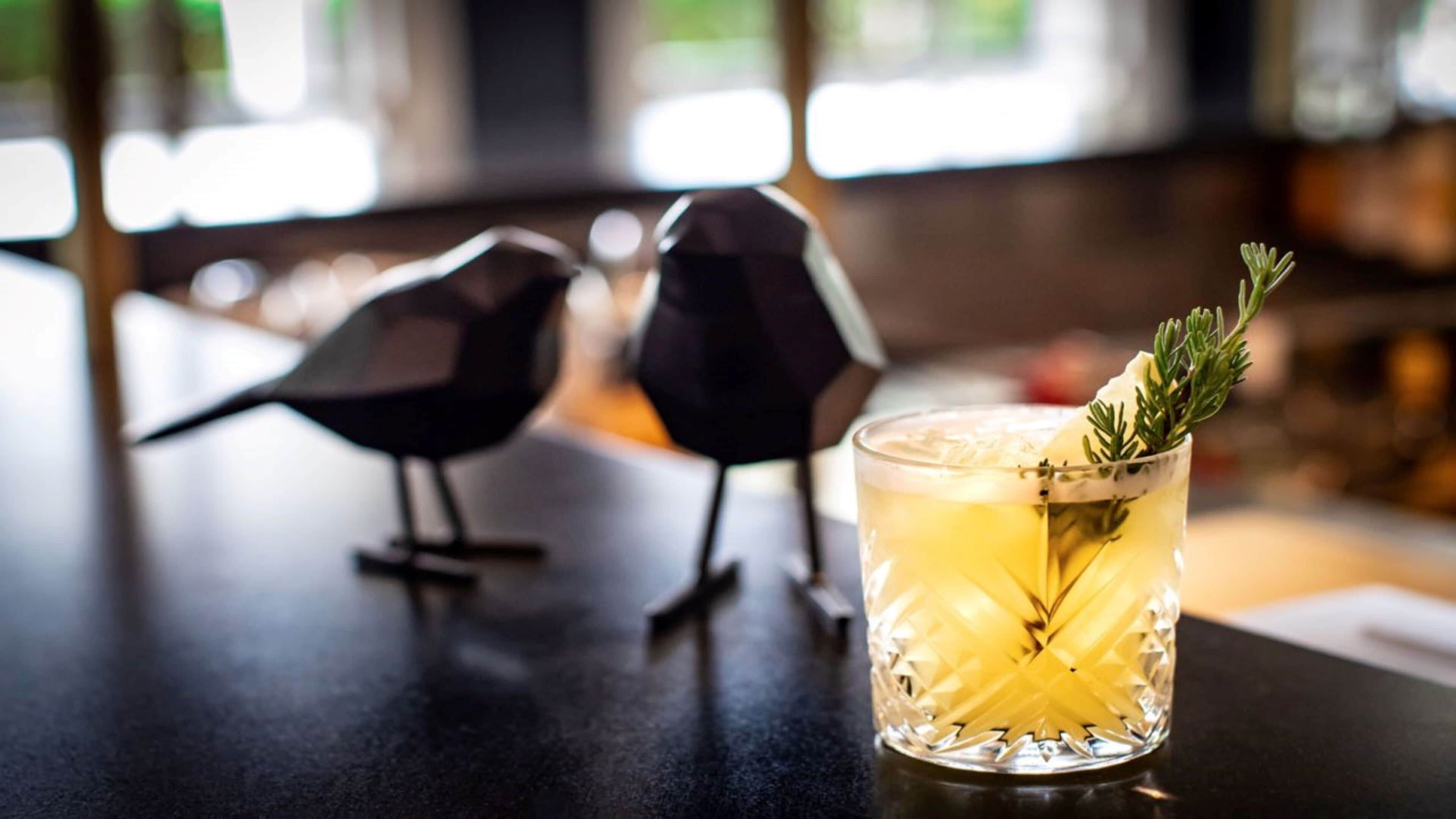 A bar setting with a cocktail glass garnished with a sprig of rosemary, and two decorative bird figures on the counter.