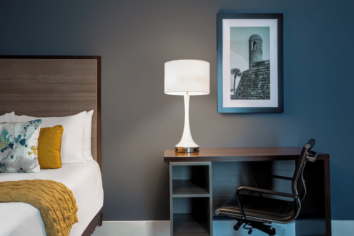 A hotel room with a bed, a desk with a lamp, a chair, and a framed picture on the wall.