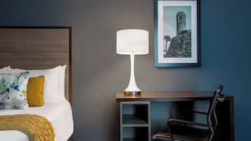 Ett hotellrum med en säng, ett skrivbord med en lampa, en stol och en inramad tavla på väggen.